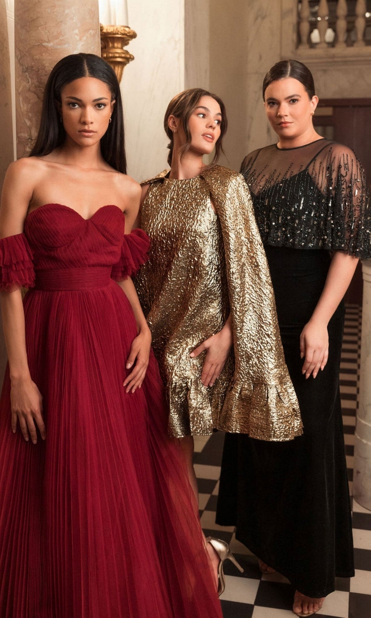 Three women pose indoors, one wearing the Mac Duggal 9228 Long Plus-Size Formal Dress in black velvet with a v-neckline; another is in an embellished sheer cape. They stand on a black-and-white checkered floor with marble columns behind them.